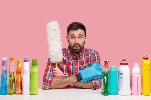 Zdjęcie zdumionego nieogolonego Europejczyka sprzątacza w kraciastej koszuli, trzyma białą szczotkę, otoczony butelkami z detergentem