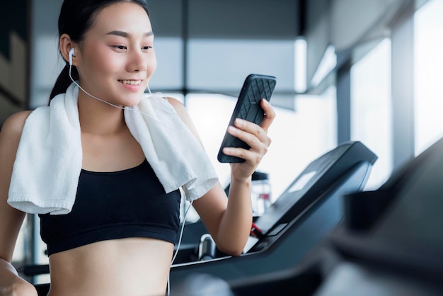 Zdrowa azjatycka kobieta w stroju sportowym odpoczywa i cieszy się piosenką ze smartfona po treningu na siłowni