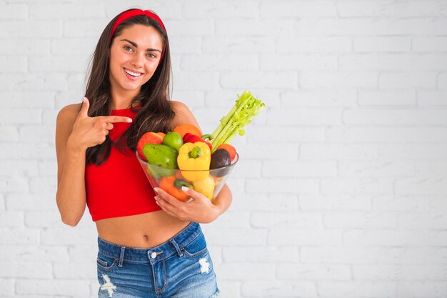 Zdrowa młoda kobieta wskazuje przy pucharem z świeżymi warzywami i owoc