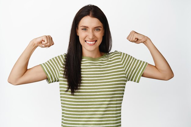Zdrowa młoda kobieta z wysportowanym ciałem napinającym bicepsy pokazujące mięśnie ramion i uśmiechniętą zadowoloną stojącą na białym tle