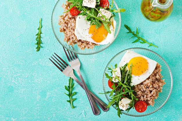 Zdrowe śniadanie z jajkiem, serem feta, rukolą, pomidorami i kaszą gryczaną na jasnym tle. Odpowiednie odżywianie. Menu dietetyczne. Leżał płasko. Widok z góry