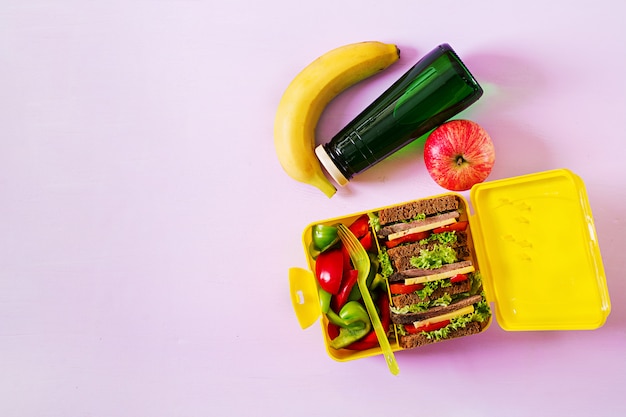 Zdrowe szkolne pudełko na lunch z kanapką wołową i świeżymi warzywami, butelką wody i owocami na różowym stole. Widok z góry. Leżał płasko