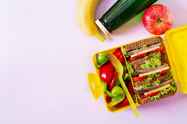 Zdrowe szkolne pudełko na lunch z kanapką wołową i świeżymi warzywami, butelką wody i owocami na różowym stole. Widok z góry. Leżał płasko