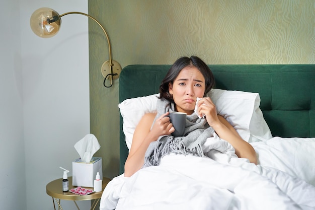 Bezpłatne zdjęcie zdrowie i ludzie, azjatycka dziewczyna leżąca w łóżku, chora, kichająca i pijąca gorącą herbatę, łapie przeziębienie