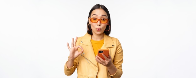 Zdumiona Azjatka w okularach przeciwsłonecznych pokazująca znak OK, trzymająca smartfona, wyglądająca na pod wrażeniem, polecając coś stojącego na białym tle