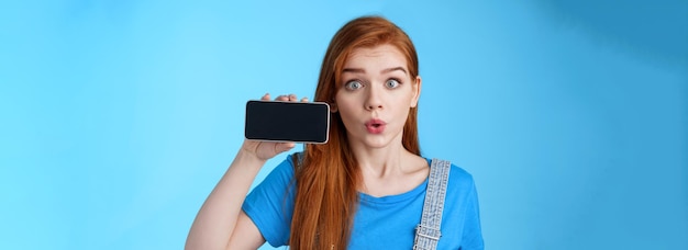 Bezpłatne zdjęcie zdumiona, zafascynowana, podekscytowana ruda kobieta pokazuje ekran smartfona poziomo składając usta, gwiżdżąc am