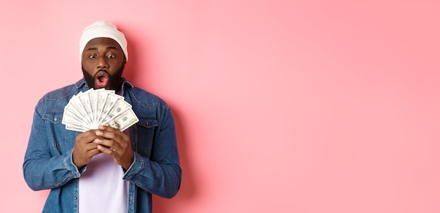 Zdumiony Afroamerykanin Otrzymuje Nagrodę Pieniężną, Pokazując Pieniądze I Wpatrując Się Z Podziwem, Stojąc Nad Różowym Ba