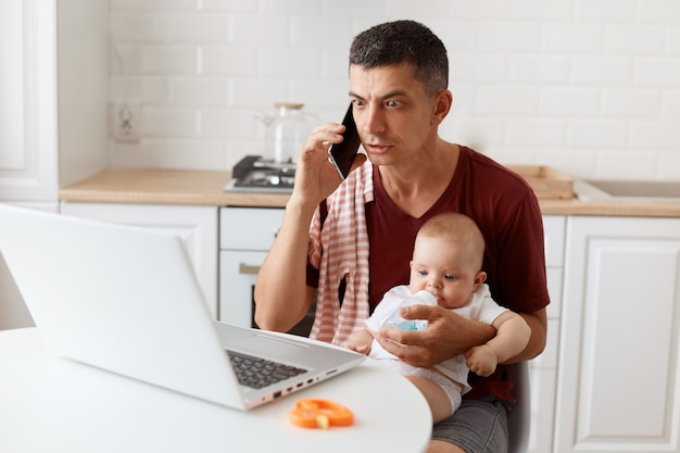 Zdumiony mężczyzna ubrany w bordową, casualową koszulkę z ręcznikiem na ramieniu, patrzy na laptopa wielkimi zszokowanymi oczami, opiekuje się dzieckiem i pracuje online z domu.