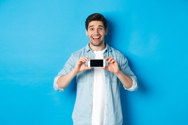 Zdumiony uśmiechnięty mężczyzna pokazujący ekran smartfona, internetową ofertę promocyjną, stojący na niebieskim tle
