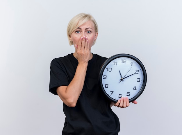 Bezpłatne zdjęcie zdziwiona blond słowiańska kobieta w średnim wieku, trzymając zegar trzymając rękę na ustach, patrząc na kamery na białym tle na białym tle z miejsca na kopię