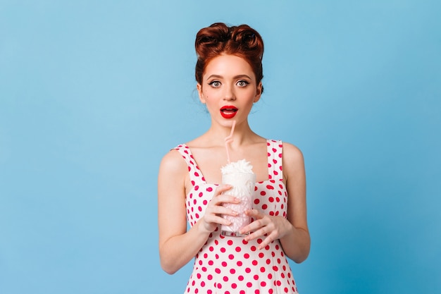 Zdziwiona dziewczyna z czerwonymi ustami pije koktajl mleczny. Studio strzałów emocjonalnej młodej damy w sukience w kropki, stojącej na niebieskiej przestrzeni.