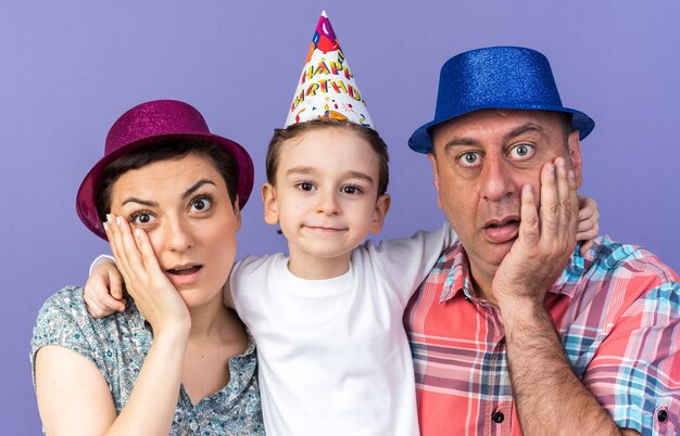 zdziwiona matka i ojciec w imprezowych czapkach kładą ręce na twarzy stojąc z synem odizolowanym na fioletowej ścianie z miejscem na kopię