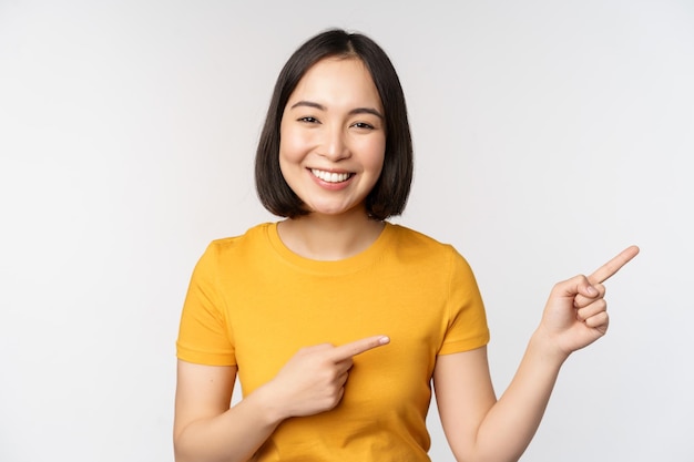 Zdziwiona młoda azjatycka kobieta pokazująca reklamę na bok, wskazując palcami na tekst promocyjny logo marki stojące szczęśliwie na białym tle