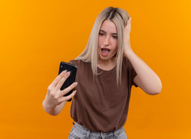 Zdziwiona młoda blondynka trzyma telefon komórkowy, patrząc na to ręką na głowie na odizolowanej pomarańczowej ścianie z miejsca na kopię