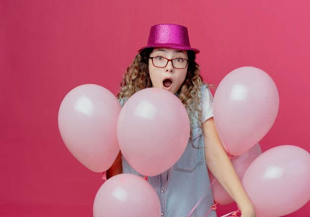 Zdziwiona młoda dziewczyna w okularach i różowym kapeluszu stojącym wśród balonów na białym tle na różowej ścianie