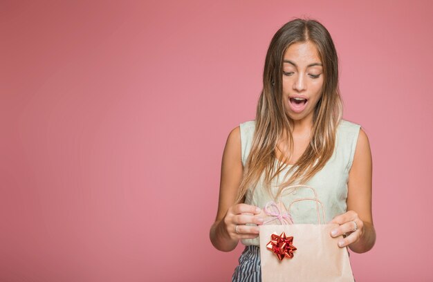 Zdziwiona młoda kobieta bierze prezent od torba na zakupy przeciw barwionemu tłu