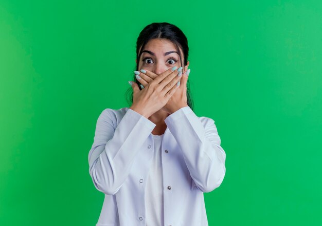 Zdziwiona młoda kobieta lekarz ubrana w szlafrok medyczny obejmujący usta rękami odizolowanymi na zielonej ścianie z miejsca na kopię