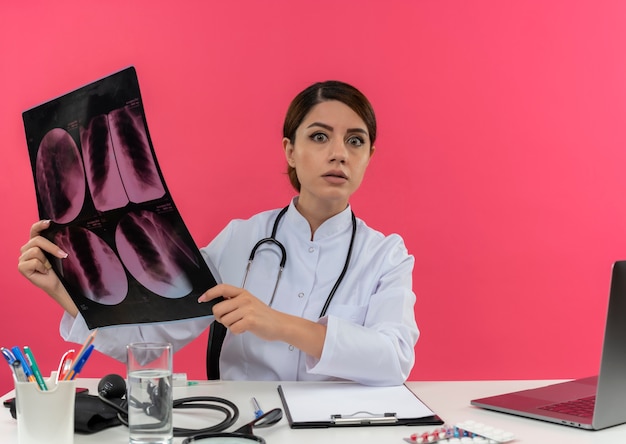 Zdziwiona młoda kobieta lekarz ubrana w szlafrok medyczny ze stetoskopem siedząca przy biurku pracuje na komputerze z narzędziami medycznymi trzymającymi prześwietlenie z miejscem na kopię