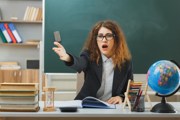 zdziwione punkty z boku młoda nauczycielka w okularach siedząca przy biurku ze szkolnymi narzędziami w klasie
