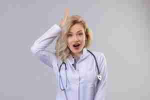 Bezpłatne zdjęcie zdziwiony lekarz młoda dziewczyna ubrana w stetoskop w sukni medycznej położyć rękę na głowie na białym tle