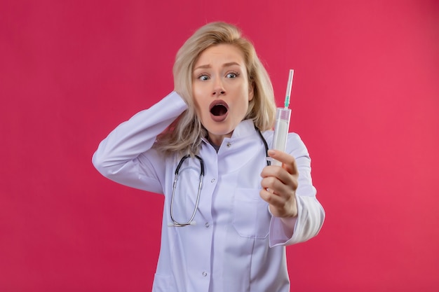Zdziwiony lekarz młoda dziewczyna ubrana w stetoskop w sukni medycznej trzymając strzykawkę chwycił głowę na czerwonym backgroung