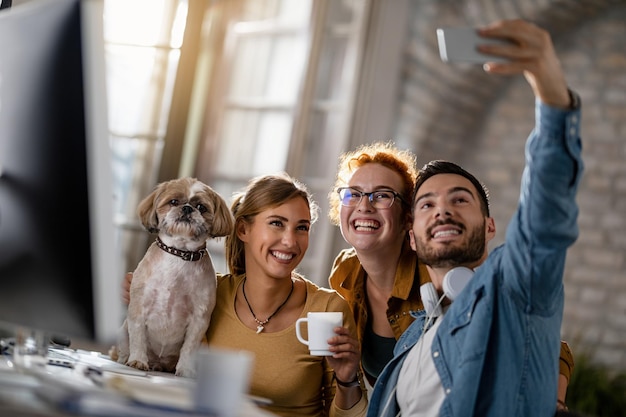 Zespół młodych, szczęśliwych ludzi biznesu z psem bawiących się podczas robienia selfie w biurze