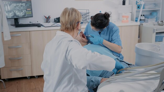 Zespół specjalistów wykonujący zabieg wiertniczy u pacjenta z bólem zęba przy użyciu narzędzi stomatologicznych w gabinecie stomatologicznym. medyk i asystentka przeprowadzająca badanie próchnicy, lecząca cierpiącego mężczyznę.