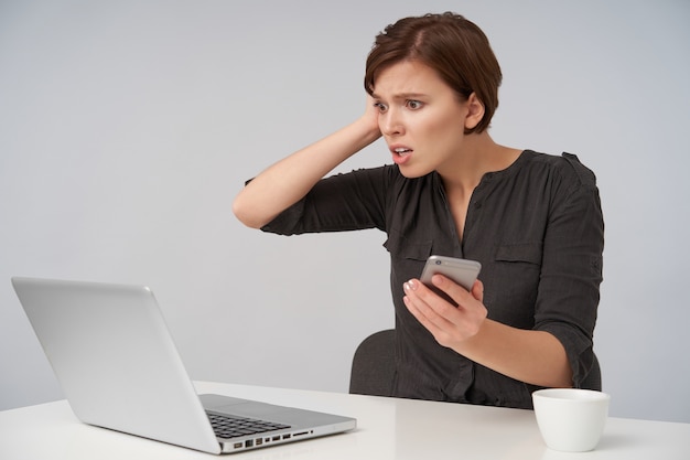 Zestresowana młoda ładna brązowowłosa kobieta z krótką modną fryzurą, trzymająca podniesioną dłoń na głowie i zaokrąglone oczy, patrząc zdumiony na ekran swojego laptopa