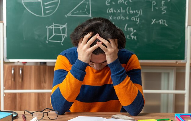 Zestresowany młody nauczyciel geometrii kaukaskiej w okularach, siedzący przy biurku z szkolnymi narzędziami w klasie, trzymając ręce na głowie