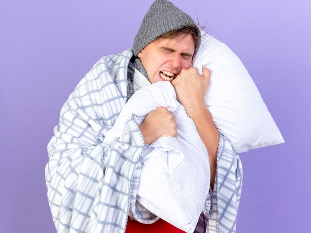 Zestresowany młody przystojny blondyn chory w czapce zimowej i szaliku owiniętym w kratę, trzymając poduszkę krzyczącą z zamkniętymi oczami odizolowanymi na fioletowym tle