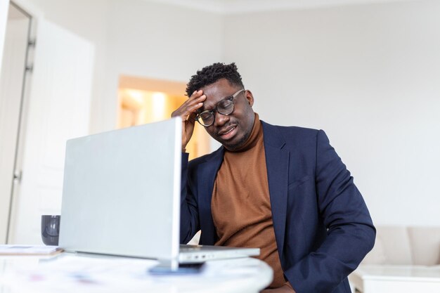 Zestresowany zmęczony Afroamerykanin dotyka skroni cierpiący na ból głowy po długich godzinach pracy przepracowany przytłoczony biznesmen siedzący przy biurku źle się czuje