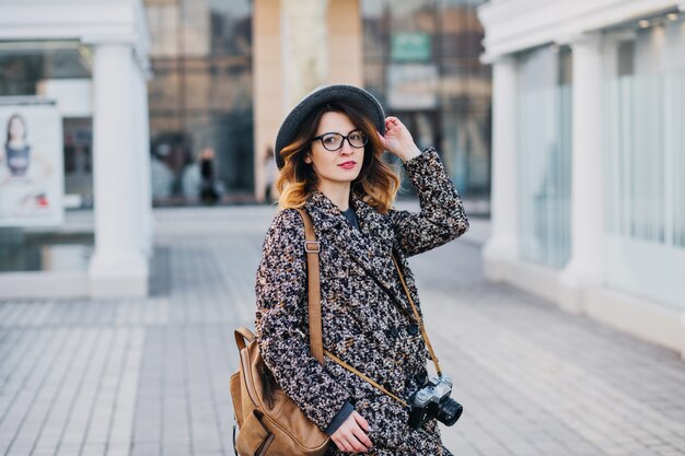 Zewnątrz portret eleganckiej młodej damy z brązowym plecakiem na sobie płaszcz i kapelusz. Atrakcyjna kobieta mówi zabawę z kręconymi włosami.