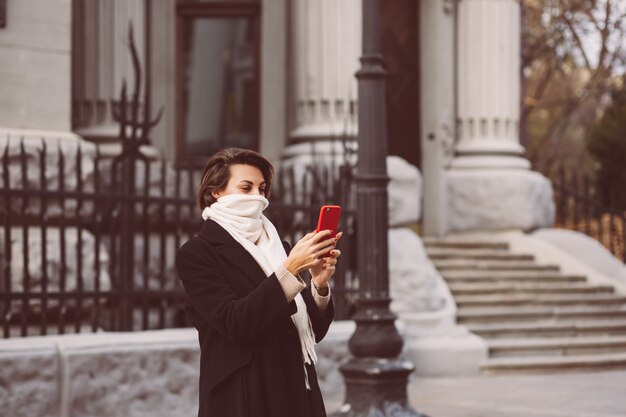Zewnątrz portret kobiety w czarnym płaszczu zimowym i białym szalikiem na ulicy, trzymając telefon komórkowy.