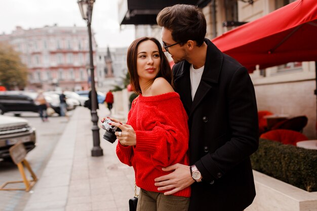 Zewnątrz portret modnej eleganckiej pary zakochanych spaceru po ulicy podczas randki lub wakacji. Brunetka dama w czerwonym swetrze robienie zdjęć aparatem.