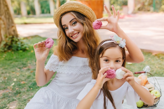 Zewnątrz portret wesoły młoda kobieta w sukienka vintage i radosna dziewczyna ze wstążką w ciemnych włosach pozowanie na charakter. Szczęśliwa matka i córka trzymając smaczne ciasteczka w parku.