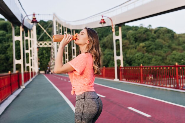 Zgrabna pani pijąca wodę podczas maratonu. Zewnątrz portret szczęśliwy biegacz kobiet stojących na torze żużlowym.