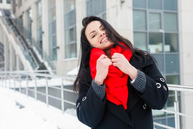 Zima, boże narodzenie, styl życia, święta i koncepcja mody - całkiem młoda uśmiechnięta kobieta w czarnym płaszczu i czerwonym szaliku pozuje w zimowym mieście. śnieg