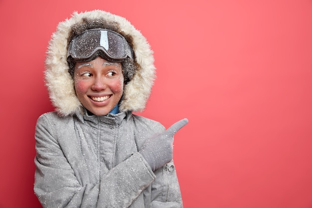 Bezpłatne zdjęcie zimowe zabawy i koncepcja jazdy na nartach. zadowolona ciemnoskóra afroamerykańska kobieta w odzieży wierzchniej wskazuje na puste miejsce, wskazując kierunek, w którym kurort cieszy się grudniowym sezonem.
