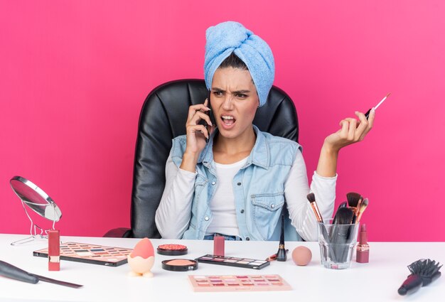 Zirytowana ładna kaukaska kobieta z zawiniętymi włosami w ręcznik, siedząca przy stole z narzędziami do makijażu rozmawiająca przez telefon i trzymająca błyszczyk na różowej ścianie z kopią przestrzeni