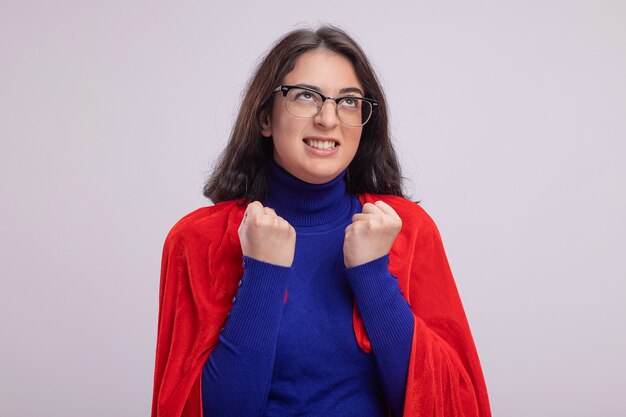 Zirytowana młoda kaukaska dziewczyna superbohatera w czerwonej pelerynie w okularach, zaciskając pięści, patrząc w górę na białym tle na białej ścianie