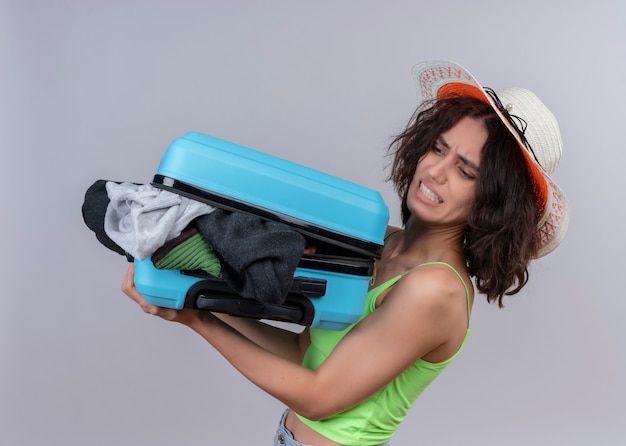 Zirytowana młoda piękna podróżna kobieta w kapeluszu i trzymając walizkę stojąc w widoku profilu na odizolowanej białej ścianie