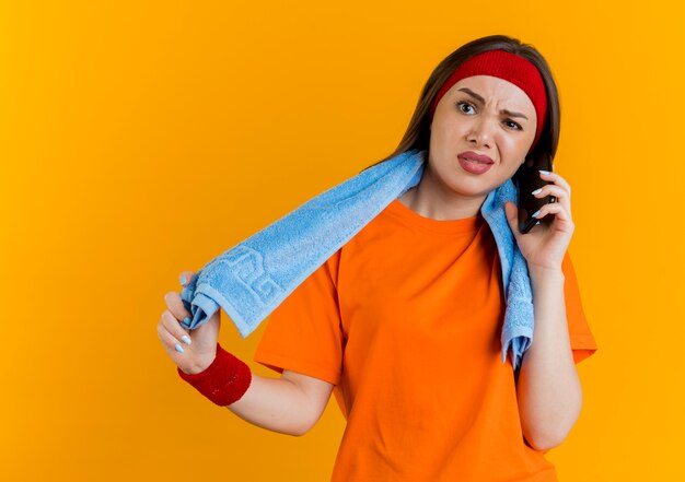 Zirytowana młoda sportowa kobieta ubrana w opaskę na głowę i opaski z ręcznikiem na szyi, chwytająca ręcznik, rozmawiająca przez telefon, patrząc prosto