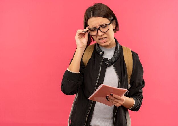 Zirytowana młoda studentka w okularach i torbie z powrotem trzymając notes i jej okulary z zamkniętymi oczami na różowym tle z miejsca na kopię