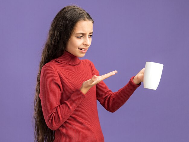 Zirytowana nastolatka trzymająca się patrząca i wskazująca ręką na filiżankę herbaty odizolowaną na fioletowej ścianie