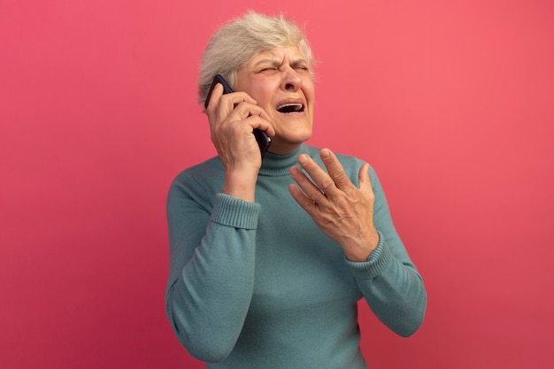 Zirytowana Stara Kobieta Ubrana W Niebieski Sweter Z Golfem Rozmawia Przez Telefon Trzymając Rękę W Powietrzu Z Zamkniętymi Oczami Odizolowanymi Na Różowej ścianie Z Kopią Przestrzeni