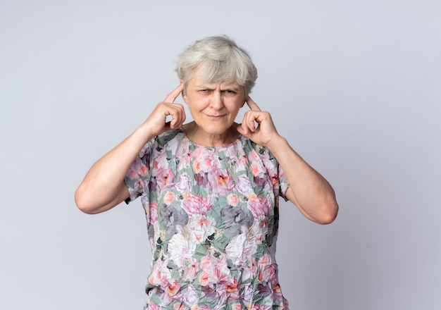 Zirytowana starsza kobieta zamyka uszy palcami na białym tle na białej ścianie