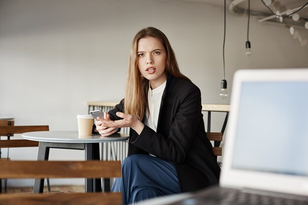 Zirytowany bizneswoman siedzieć w kawiarni z telefonem komórkowym i patrzeć na zirytowaną osobę