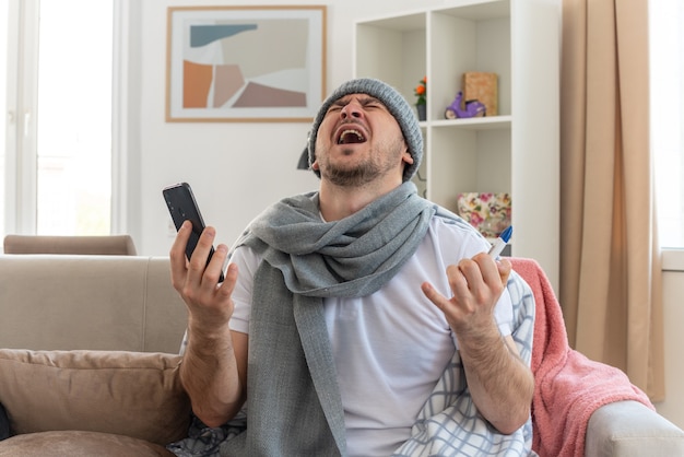 zirytowany chory mężczyzna z szalikiem na szyi w czapce zimowej trzymający termometr i telefon siedzący na kanapie w salonie