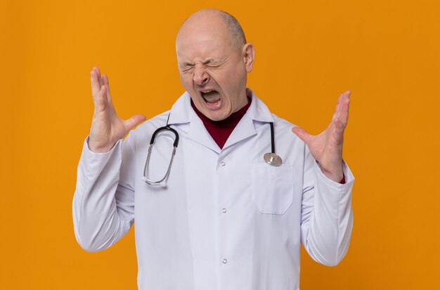 Zirytowany dorosły słowiański mężczyzna w mundurze lekarza ze stetoskopem trzymającym ręce otwarte i krzyczącym