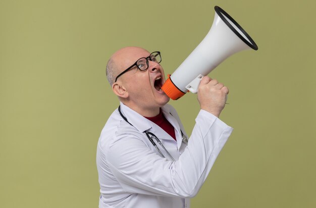 Zirytowany dorosły słowiański mężczyzna w okularach optycznych w mundurze lekarza ze stetoskopem krzyczącym do głośnika patrzącego w górę